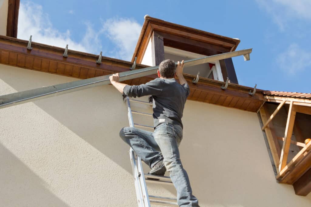Un homme sur une échelle installe une gouttière métallique sur la façade d'une maison en bois sous un ciel bleu dégagé.