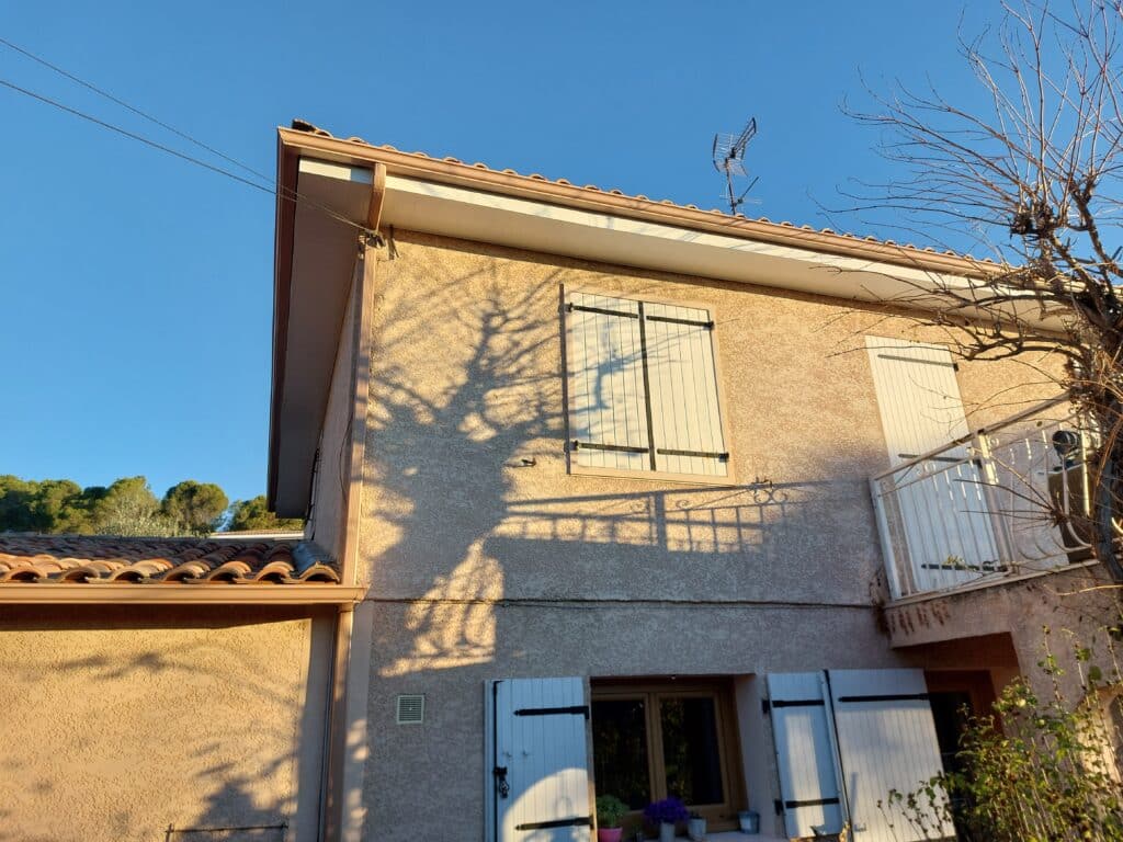 Maison méditerranéenne en stuc, façade illuminée par le soleil couchant. L'ombre d'un arbre sans feuilles se projette sur le mur et les volets blancs du premier étage.