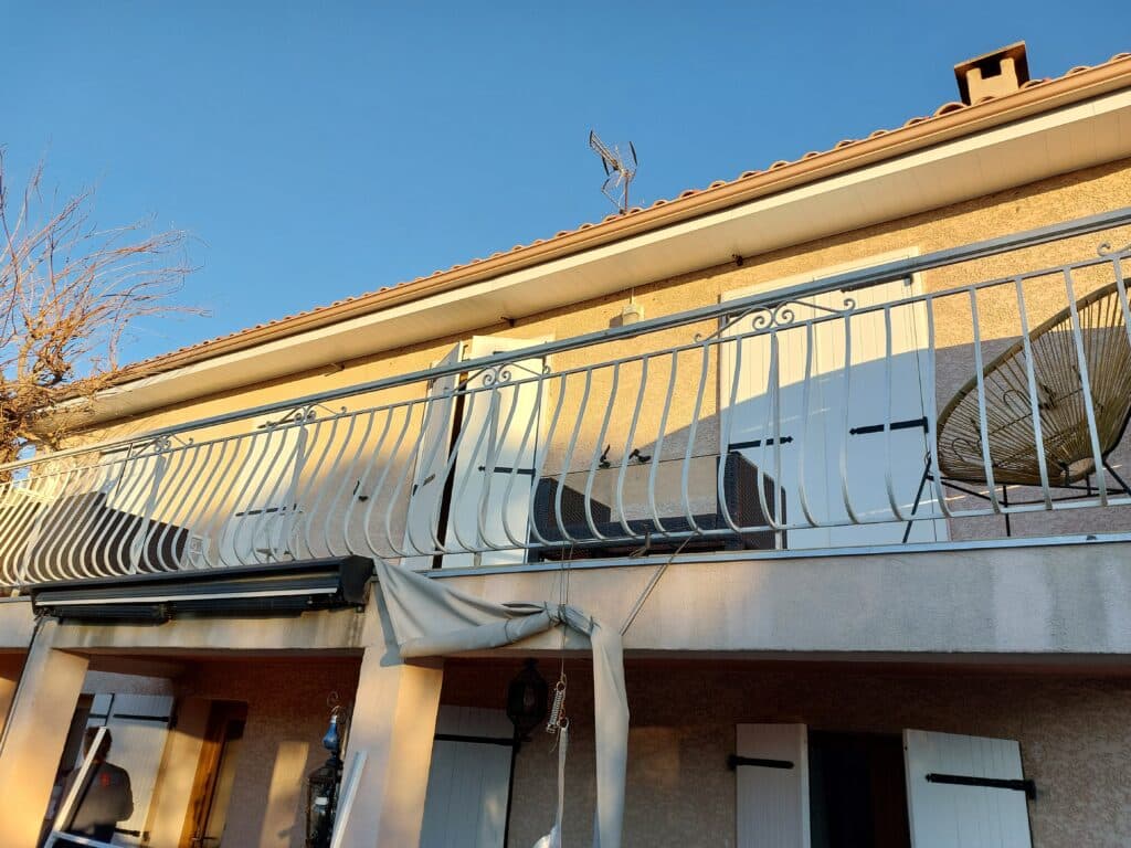 Balcon avec garde-corps blanc courbé, volets blancs et antenne TV sur une façade de maison sous un ciel bleu dégagé.
